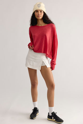 A model wearing a red long-sleeve knit top, a white asymmetrical mini skirt, a cream beanie, white socks, and black sneakers, standing against a light gray studio background.