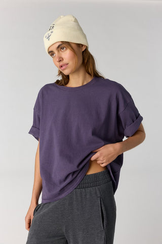 A woman wearing a loose, short-sleeve purple t-shirt with rolled cuffs, dark gray sweatpants, and a cream beanie, posing against a light gray studio background.