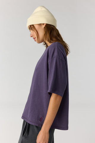 A person shown in side profile wearing an oversized short-sleeve purple t-shirt, dark gray pants, and a cream-colored beanie against a light gray studio background.