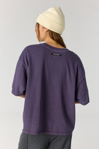 A person shown from the back wearing an oversized short-sleeve purple t-shirt, a cream-colored beanie, and dark gray pants against a light gray studio background.