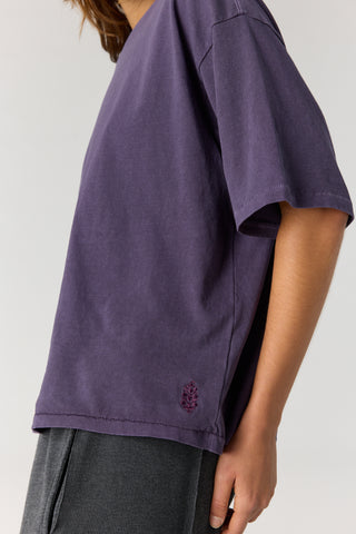 A close-up side view of a person wearing an oversized short-sleeve t-shirt in washed purple. The shirt features a small tonal embroidered logo near the lower left hem and is paired with dark gray corduroy pants against a light gray studio background.