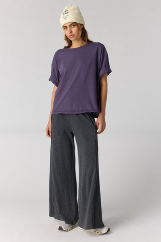 A model wearing a loose-fitting purple short-sleeve t-shirt, dark gray wide-leg pants, a cream beanie, and leopard print shoes, standing against a light gray studio background.