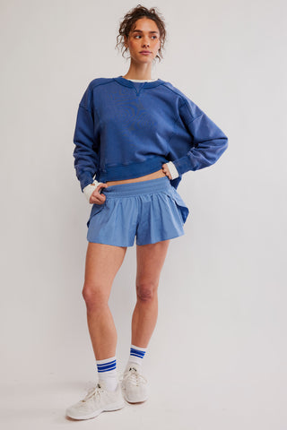 A model wearing a blue crewneck sweatshirt, matching blue shorts, white socks with blue stripes, and white sneakers, standing against a light gray studio background.