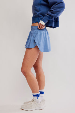 A model wearing a light blue athletic wrap mini skirt, a blue sweatshirt, white socks with blue stripes, and white sneakers, standing against a light gray studio background.