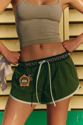A close-up lifestyle shot of a model wearing dark green athletic shorts with a black elastic waistband featuring the white text 'MOVEMENT' and a white drawstring. The model is also wearing a gray ribbed crop top, standing against a yellow wooden slatted wall.