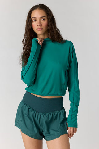 A woman modeling a teal green long-sleeve cropped athletic top and matching green shorts against a light gray studio background.