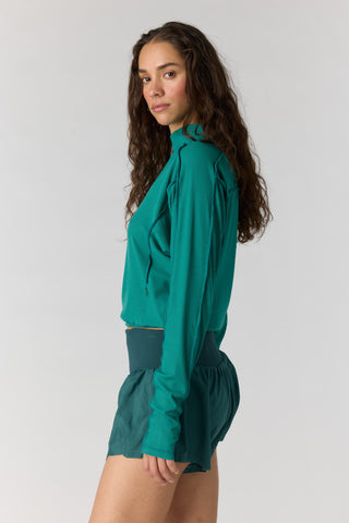 A woman modeling a teal green long-sleeve blouse and matching high-waisted shorts against a light gray studio background.