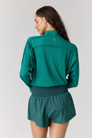 A woman shown from the back wearing a teal-green athletic zip-up jacket and matching green shorts against a light gray studio background.