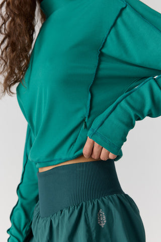 A close-up of a person wearing a teal-green ribbed long-sleeve crop top and matching high-waisted athletic bottoms against a light gray studio background.