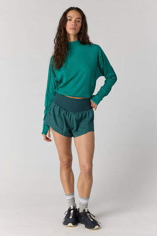 A woman modeling a matching athletic set consisting of a teal green long-sleeve mock-neck crop top and dark teal green running shorts, paired with gray socks and black sneakers, against a light gray studio background.
