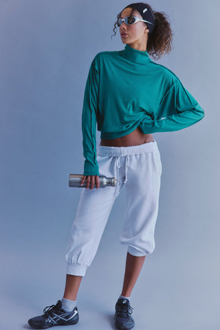 A woman modeling a teal green long-sleeve mock neck crop top and white jogger sweatpants, holding a silver water bottle against a solid blue-gray studio background.