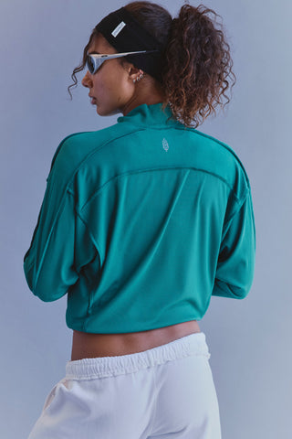 A woman wearing a teal green athletic cropped long-sleeve top and white sweatpants, shown from the back against a solid gray studio background.