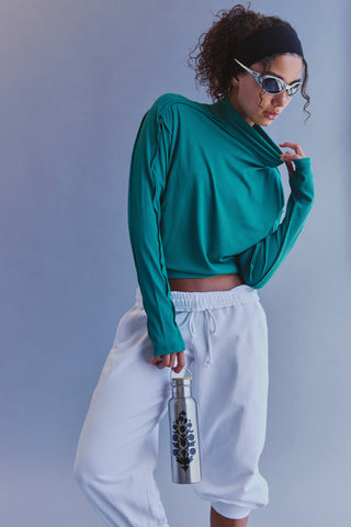 A woman wearing a teal long-sleeve mock neck crop top and white sweatpants, holding a silver water bottle with a black floral design against a solid blue-gray studio background.
