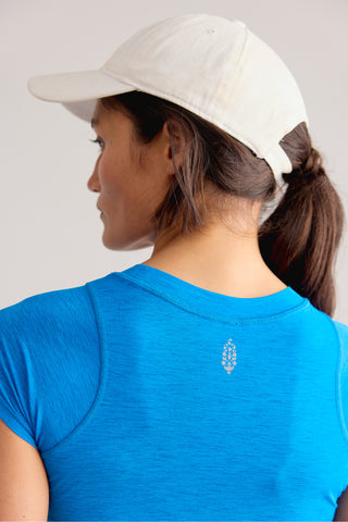 A woman shown from the back wearing a bright blue athletic t-shirt and a white baseball cap, with a small white embroidered logo on the center of the shirt.