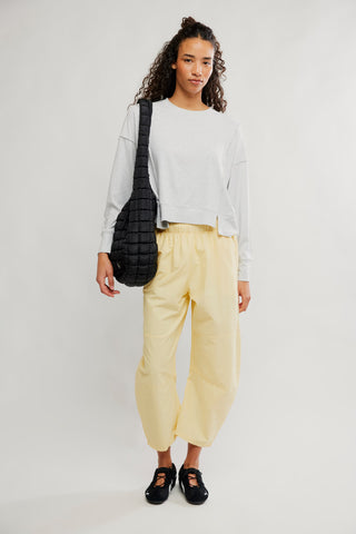 A model stands against a light gray studio background wearing a white cropped sweatshirt, pale yellow cropped pants, black shoes, and carrying a black quilted shoulder bag.
