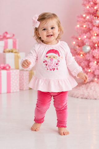 A toddler girl wearing a light pink long-sleeve ruffle-hem top with a Santa-clad graphic and the words 'let it Snow' in pink, paired with bright pink leggings, standing in a festive pink holiday setting with a decorated tree and wrapped gifts.