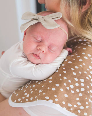 Newborn baby in a white outfit with a bow, held by a person wearing a patterned dress.