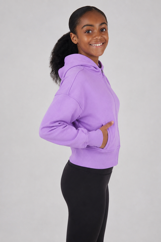 A young girl with dark curly hair in a ponytail, wearing a lavender purple cropped hoodie and black leggings, posing in profile against a light gray studio background.
