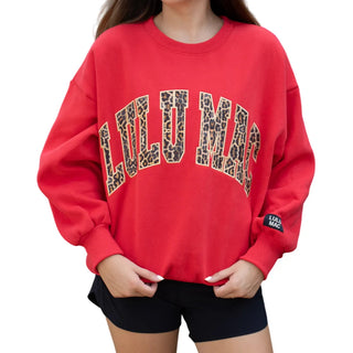 A woman wearing a red crewneck sweatshirt with 'LULU MAC' stitched in leopard print across the chest, paired with black shorts, against a plain white background.