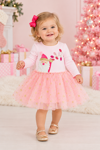 A toddler girl wearing a pink long-sleeve top with a gnome graphic and a matching pink tulle tutu skirt, standing in a festive holiday room with a decorated pink tree and wrapped gifts.