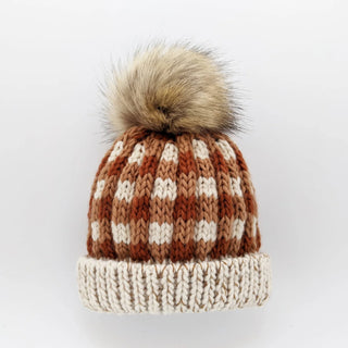 A knit pom-pom beanie hat featuring a brown and cream checkerboard pattern on the crown and a solid cream ribbed cuff, topped with a fluffy faux-fur pom-pom.