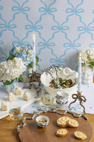 Decorative table setting with flowers, candles, and food on a tablecloth against a patterned wall.