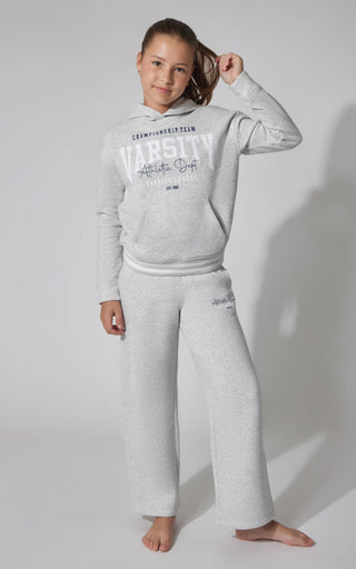 A young girl modeling a light gray heathered two-piece loungewear set, consisting of a long-sleeve hoodie with a 'VARSITY' graphic on the chest and matching wide-leg sweatpants, against a plain light gray studio background.