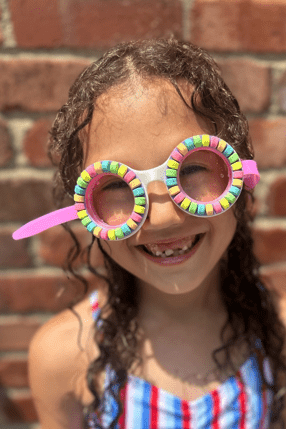Child wearing colorful swim goggles and a striped swimsuit in front of a brick wall.