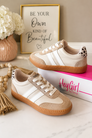 A pair of low-top sneakers styled on a marble surface with a pink and white box, a framed quote, and flowers in the background. The shoes are cream and beige with white side stripes, tan suede overlays, and gum soles.
