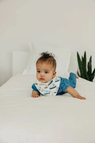 Baby wearing a bib and blue outfit lying on a white bed with a plant in the background