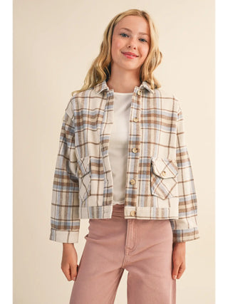 A young woman modeling a cropped, long-sleeve plaid button-up shirt jacket in beige, brown, and light blue, worn over a white t-shirt and pink pants against a solid beige background.
