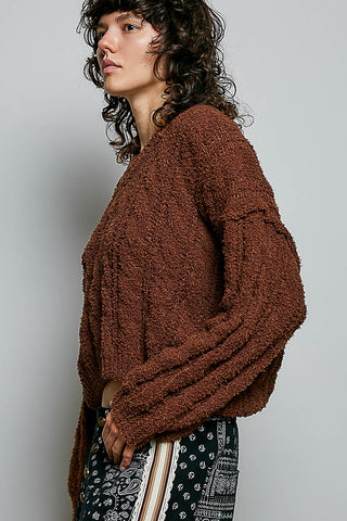A woman wearing a brown, chunky, textured boucle knit sweater and patterned pants, shown in a side-profile view against a plain light gray studio background.