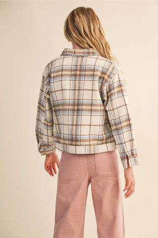A woman shown from the back wearing a cropped plaid flannel shirt jacket in beige, brown, and light blue, paired with dusty pink pants, against a solid light beige background.
