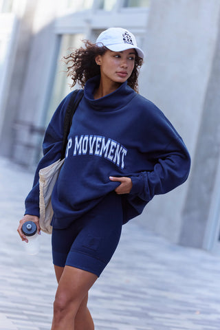 A woman walking outdoors wearing an oversized navy blue sweatshirt with 'FP MOVEMENT' printed in white across the chest, matching navy biker shorts, a white baseball cap, and carrying a beige shoulder bag.