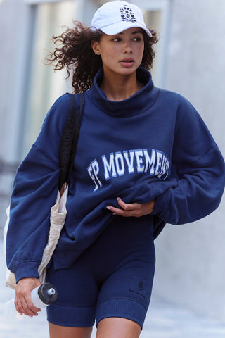 A woman wearing a navy blue sweatshirt and matching shorts with 'FP MOVEMENTS' printed in white across the chest, paired with a white baseball cap and a beige shoulder bag.