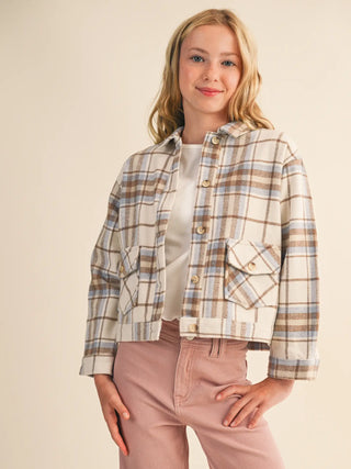 A young woman modeling a cropped plaid button-up jacket with a hood, worn over a white top and pink pants, against a solid beige background.