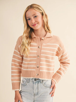 A young blonde woman wearing a cropped, long-sleeve knit cardigan with horizontal peach and cream stripes, paired with light-wash denim jeans, against a solid beige studio background.