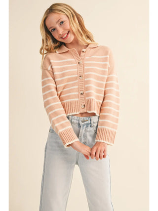 A young girl with blonde hair wearing a cropped, long-sleeve knit cardigan with horizontal peach and cream stripes, paired with light-wash jeans, against a solid beige background.