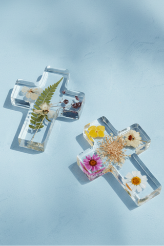 Two cross-shaped resin paperweights with pressed flowers on a light blue background
