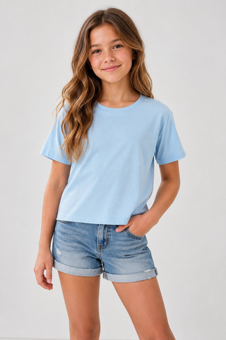 A young girl with long wavy brown hair wearing a light blue short-sleeve crewneck t-shirt and light-wash denim shorts, standing against a plain light gray studio background.