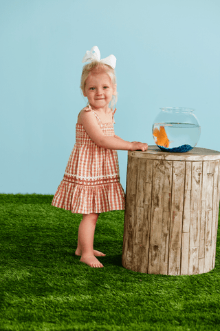 Child in a checkered dress standing next to a wooden stump with a fishbowl on a grassy area.