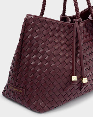 A close-up of a woven leather tote bag in deep burgundy, featuring braided shoulder straps, a drawstring detail with gold-tone hardware, and a small 'Little London' brand patch near the bottom left corner.