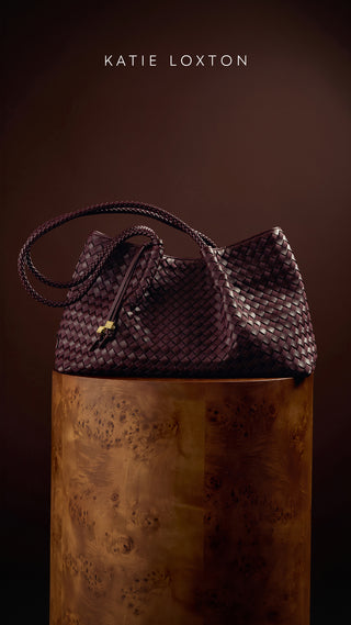 A dark woven leather handbag with braided straps and a small gold accent, displayed on a polished wooden pedestal against a dark brown background. The text 'KATIE LOXTON' appears at the top.