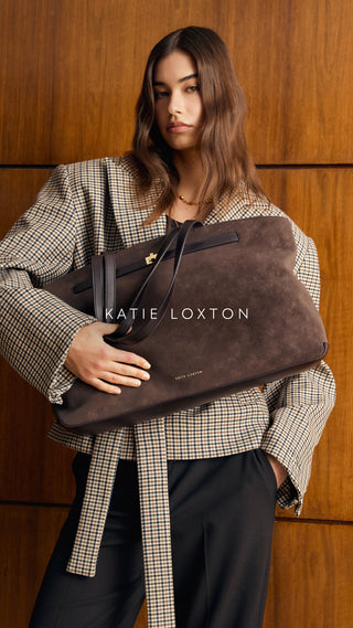 A model wearing a beige and black checkered blazer and black trousers holds a large brown suede Katie Loxton bag against a warm wood-paneled background.