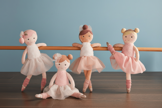Four ballerina dolls in pink outfits standing on a wooden floor against a blue wall.