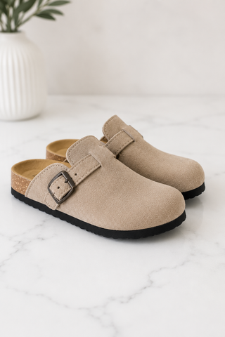 A pair of beige suede clogs with a black sole and adjustable strap, displayed on a white marble surface with a white vase in the background.