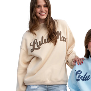 A woman wearing a cream-colored oversized sweatshirt with 'Lulu Made' printed in brown script across the chest, standing next to another person in a light blue sweatshirt against a white background.
