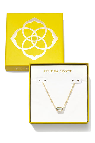 Kendra Scott jewelry set featuring a delicate gold chain necklace with a small oval pendant, displayed in an open white gift box with a yellow border. The matching yellow lid is positioned behind the box, showcasing the brand's geometric logo.