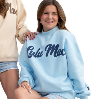 A woman wearing a light blue crewneck sweatshirt with 'Celu Mac' printed in navy script across the chest, seated against a white background with another model partially visible on the left.