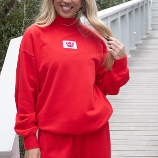 A woman wearing a bright red sweatshirt and matching sweatpants stands on a light-colored wooden walkway. The sweatshirt has a mock neck and a small white chest patch with the text 'LULU MAC' in red.